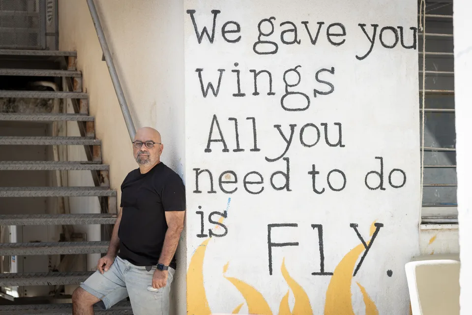 דרור שגב נשען על קיר שעליו מצויר הכיתוב: We gave you wings, all you need to do is fly