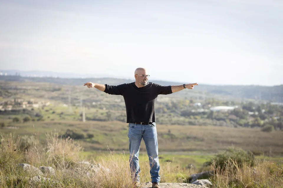 דרור שגב עומד על סלע בשדה פתוח, ידיו פרושות לצדדים כמו כנפיים
