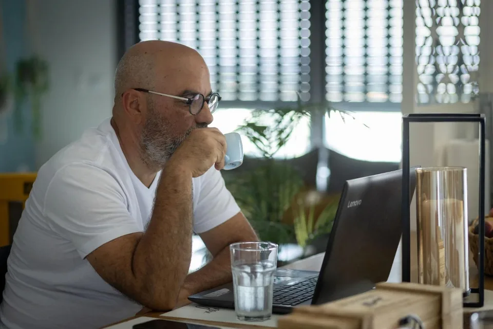 דרור שגב יושב מול המחשב הנייד עם כוס קפה, מהורהר, באור טבעי